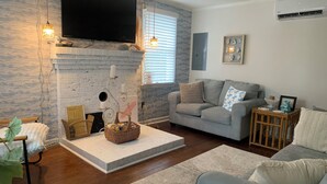 Un salon doté d’une cheminée, d’un téléviseur à écran plat fixé au-dessus, d’un canapé gris, d’une petite table d’appoint en bois et d’un panier en osier posé sur une plateforme blanche.