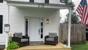 Une maison avec un porche, au numéro 210, qui comprend deux fauteuils en osier, une porte blanche et un drapeau américain.