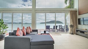 Amplia sala de estar con un sofá gris, cojines rojos y beige, y vista al mar a través de grandes ventanales.