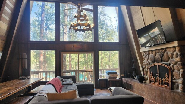 A spacious loft-style room with a large window overlooking a forest, a stone fireplace, and a flat-screen TV mounted above it. - The Cub House: Mountain Home w/ Hot Tub, Pool Table, 5 bed/4ba, Fireplace, Woods (Ruidoso)