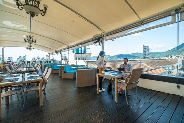 Un espace extérieur abrité, aménagé pour les repas, avec des tables et des chaises, une personne qui sert les clients, et une vue sur le paysage urbain.