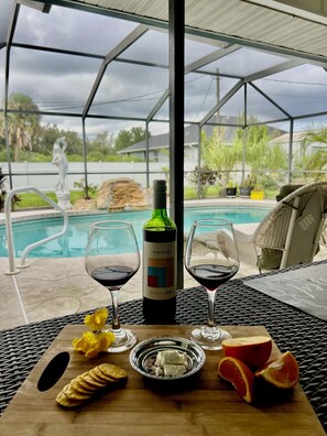 Une table placée au bord de la piscine, avec une bouteille de vin rouge, deux verres, des crackers, du fromage et des agrumes.