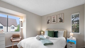 Een net opgemaakt bed met witte lakens en een groene plaid, een nachtkastje met een lamp, en een raam met uitzicht op een huis.