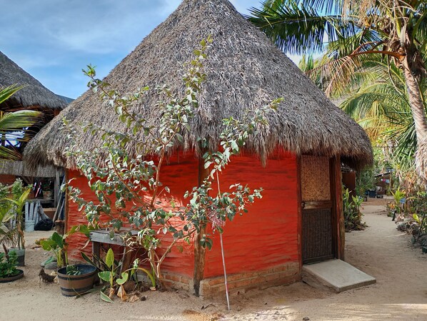 A thatched-roof hut with a red exterior, surrounded by plants and palm trees.