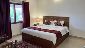A hotel room with a bed, a nightstand, a lamp, a chair, and a window with curtains. - River Meadows  (Tiruvalla)
