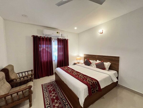 A bedroom with a bed, a chair, a rug, and a window with curtains. - River Meadows (Tiruvalla)
