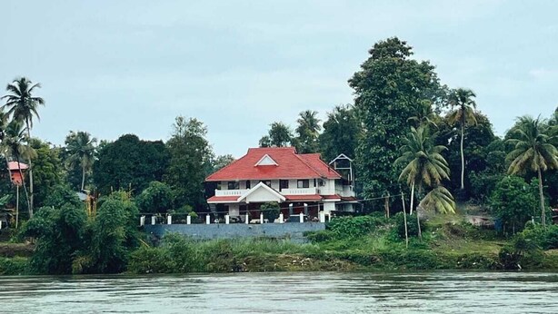 A two-story building with a red roof, surrounded by lush greenery and palm trees, located near a body of water. - River Meadows  (Tiruvalla)