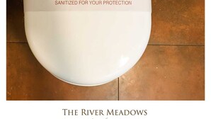 A white toilet with a label stating "SANITIZED FOR YOUR PROTECTION" and the text "THE RIVER MEADOWS luxury home stay" below it. - River Meadows  (Tiruvalla)