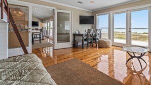 A spacious room with a wooden floor, a ceiling fan, a dining area with a table and chairs, and a large window offering a view of the outdoors. - Bay Breeze - Waterfront with Boat Dock, Helipad! (Panacea)