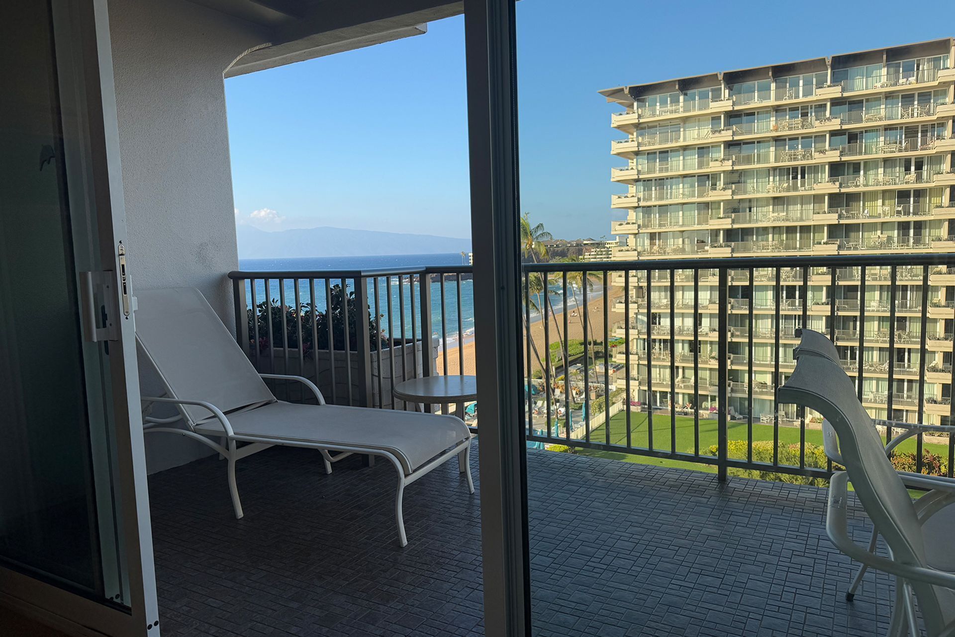 Un balcon avec deux fauteuils de jardin blancs, une petite table et une vue sur une plage et un immeuble de grande hauteur.