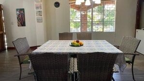 Un comedor con una mesa dispuesta para cuatro, sillas de mimbre, una bandeja de frutas, un reloj de pared y una ventana con vistas al verdor.