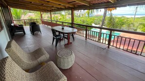 Un espacio al aire libre cubierto con una mesa y sillas de madera, pufs tejidos y vistas a una piscina y follaje.