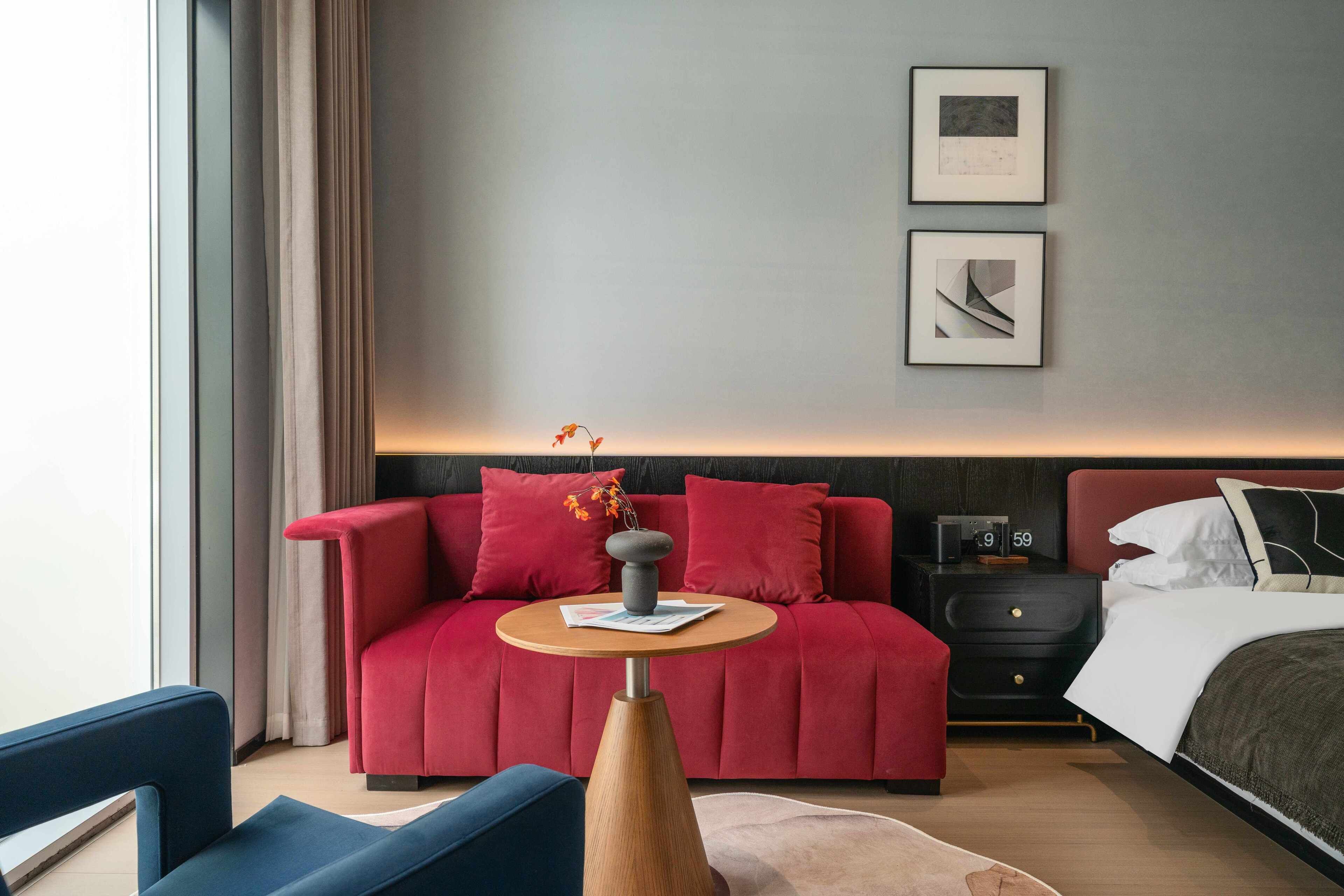 A modern hotel room with a red sofa, a small round table, a bed, and two framed pictures on the wall.