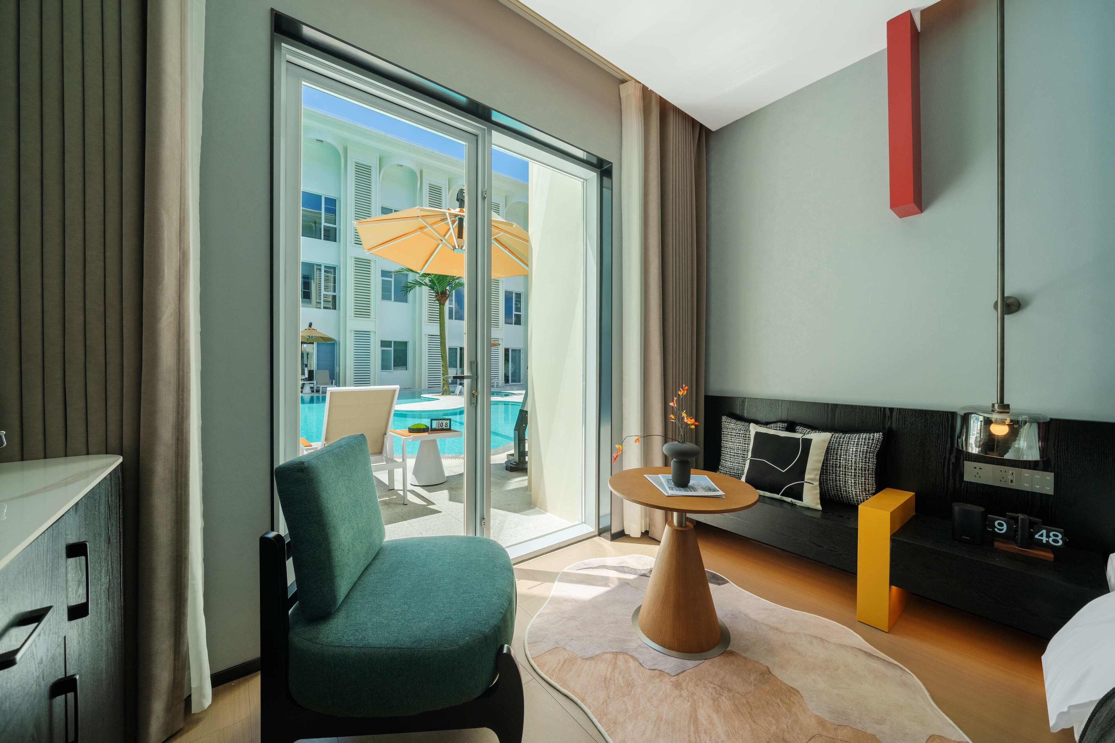 Una habitación de hotel moderna con un amplio ventanal corredero que da a una zona de piscina, un sofá con mesita auxiliar y un escritorio con silla.