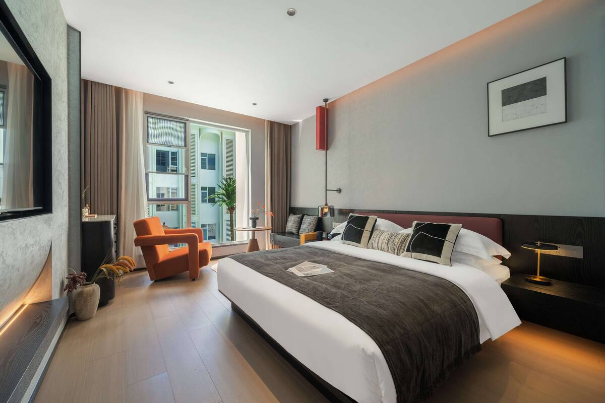 A modern hotel room with a large bed, a seating area with a chair, a bedside table, and a view of the city through large windows.