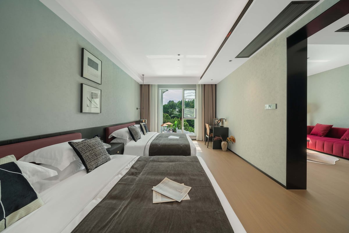 A hotel room with two beds, a red sofa, and a view of greenery outside.