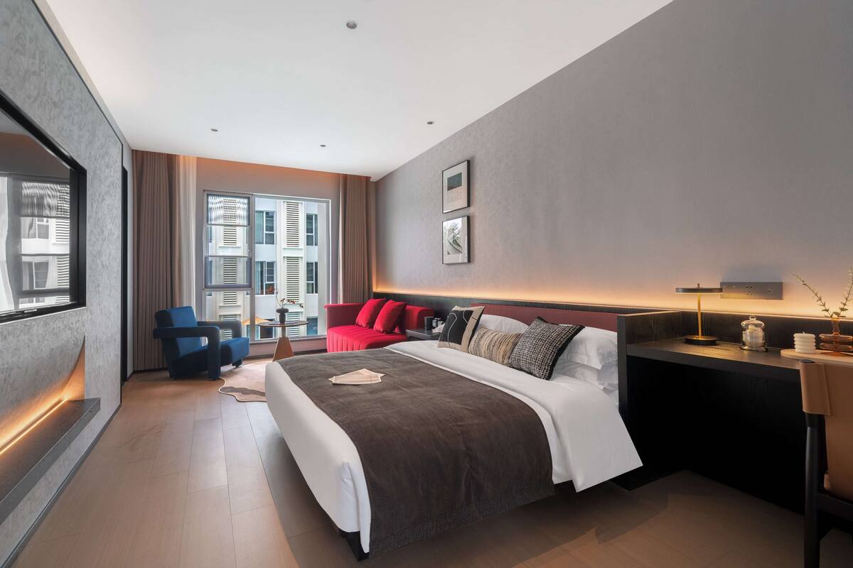 A modern hotel room with a large bed, a desk, a red sofa, and a large window offering a city view.