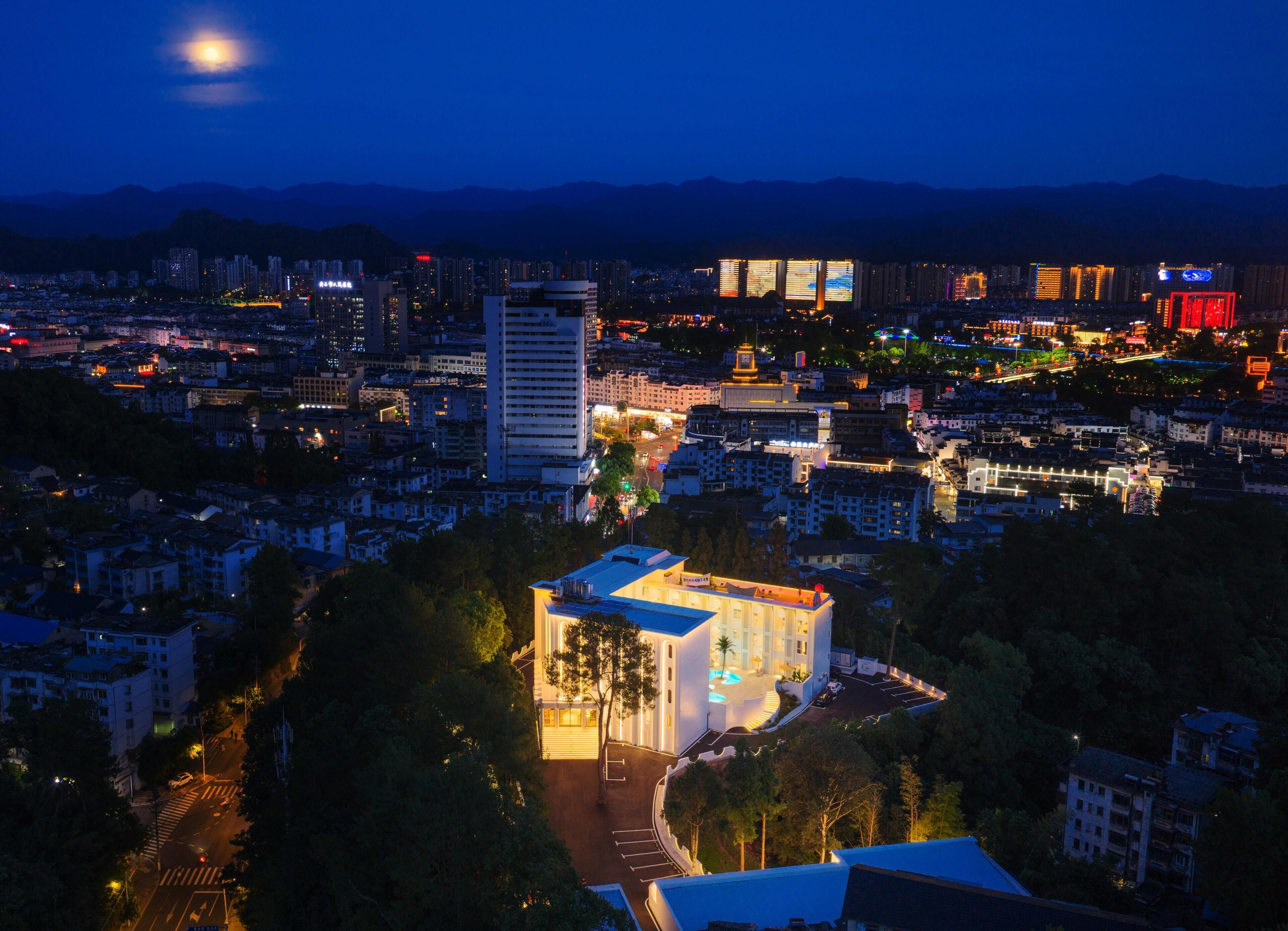 A cityscape at night with illuminated buildings, a prominent white hotel with a swimming pool, and a mountain range in the background.