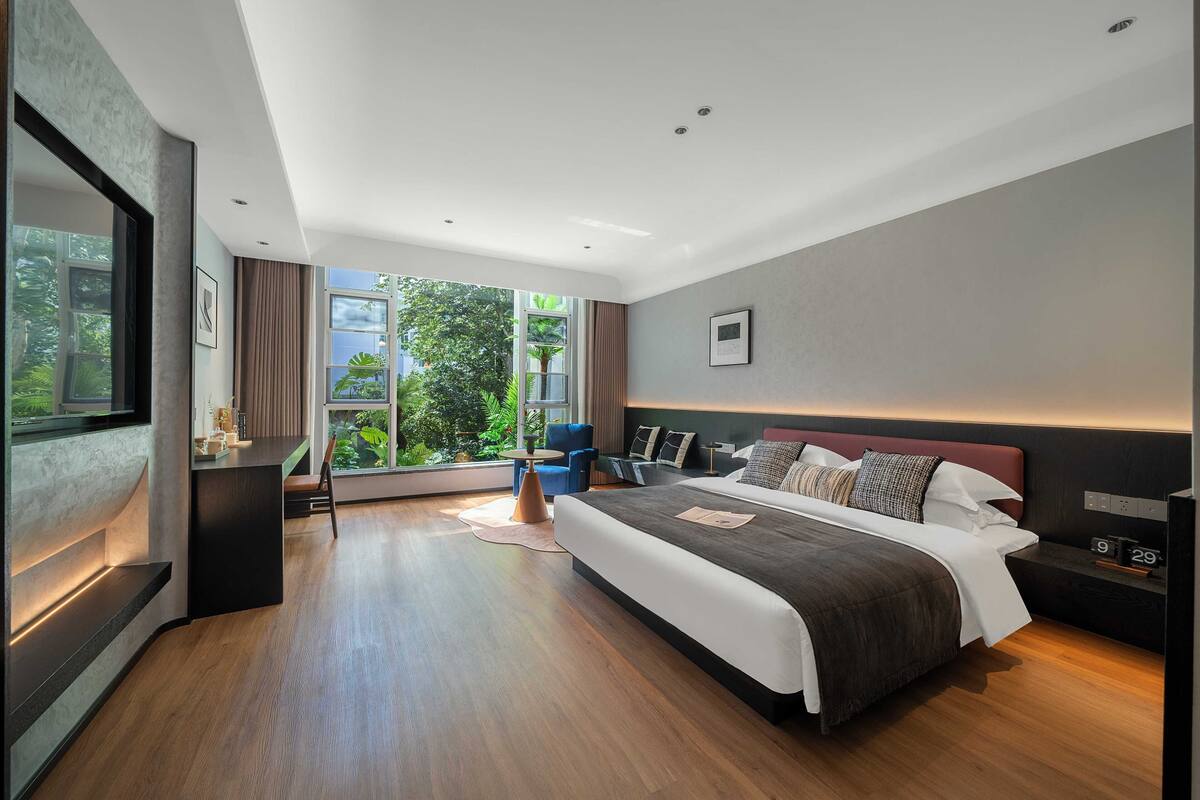 A modern bedroom with a large bed, a desk, and a view of greenery through the window.