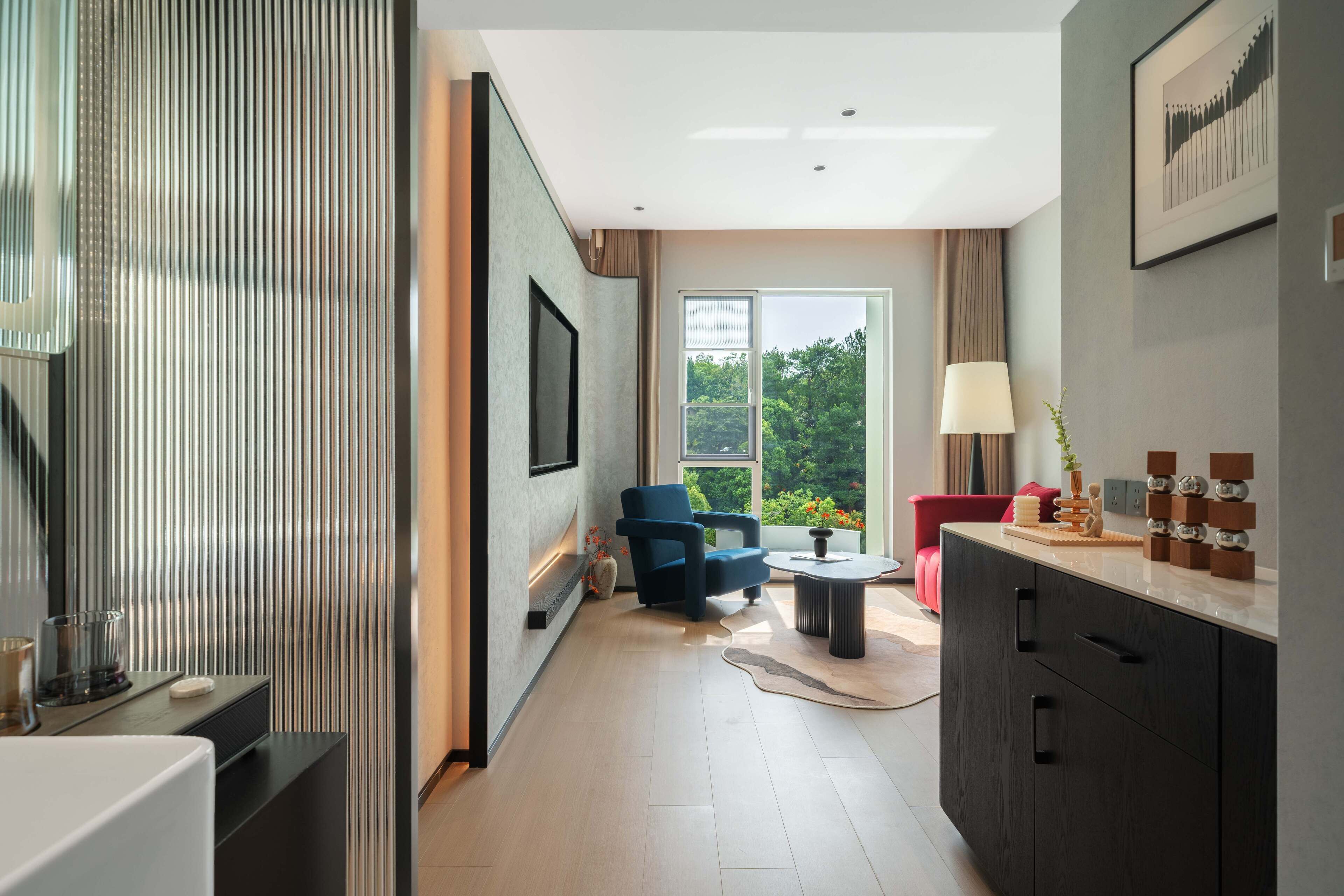 A modern hotel room with a large window, a seating area with a blue chair, a small table, and a cabinet with drawers.