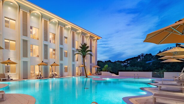 A swimming pool with lounge chairs and umbrellas, surrounded by a building with large windows and a palm tree. - MisooHi Hidden Realm Hotel (Huangshan)