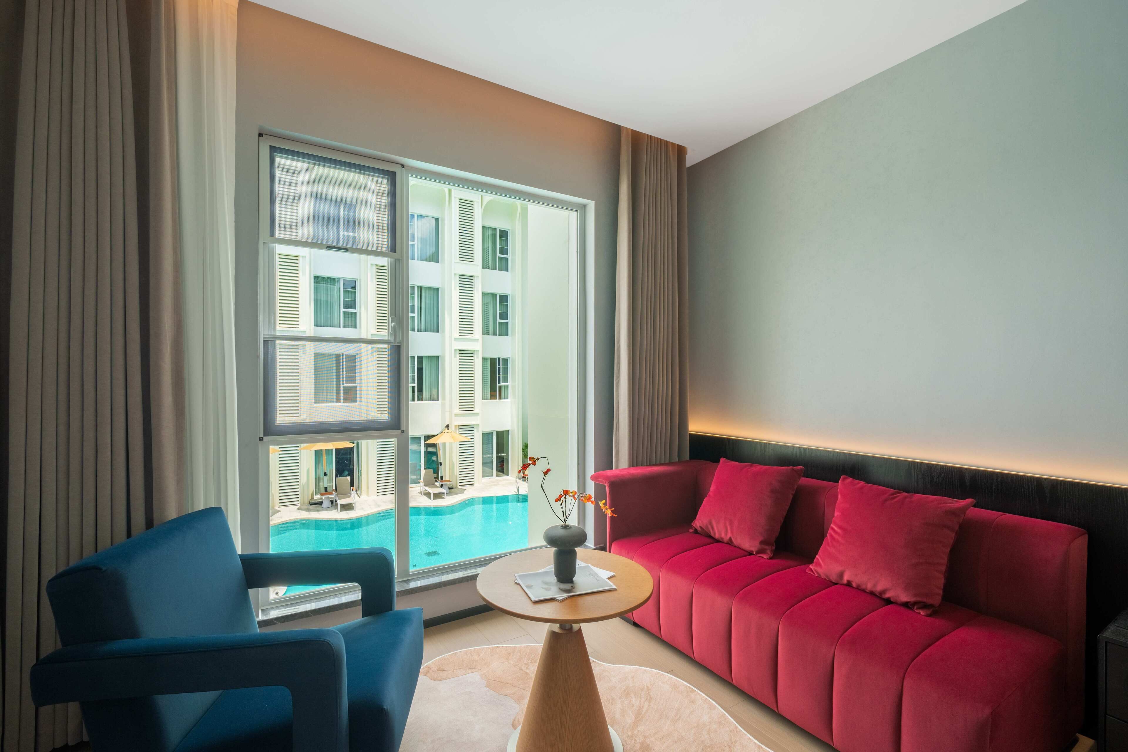 A modern hotel room with a red sofa, a blue armchair, a small round table with a vase, and a large window offering a view of a pool and buildings.