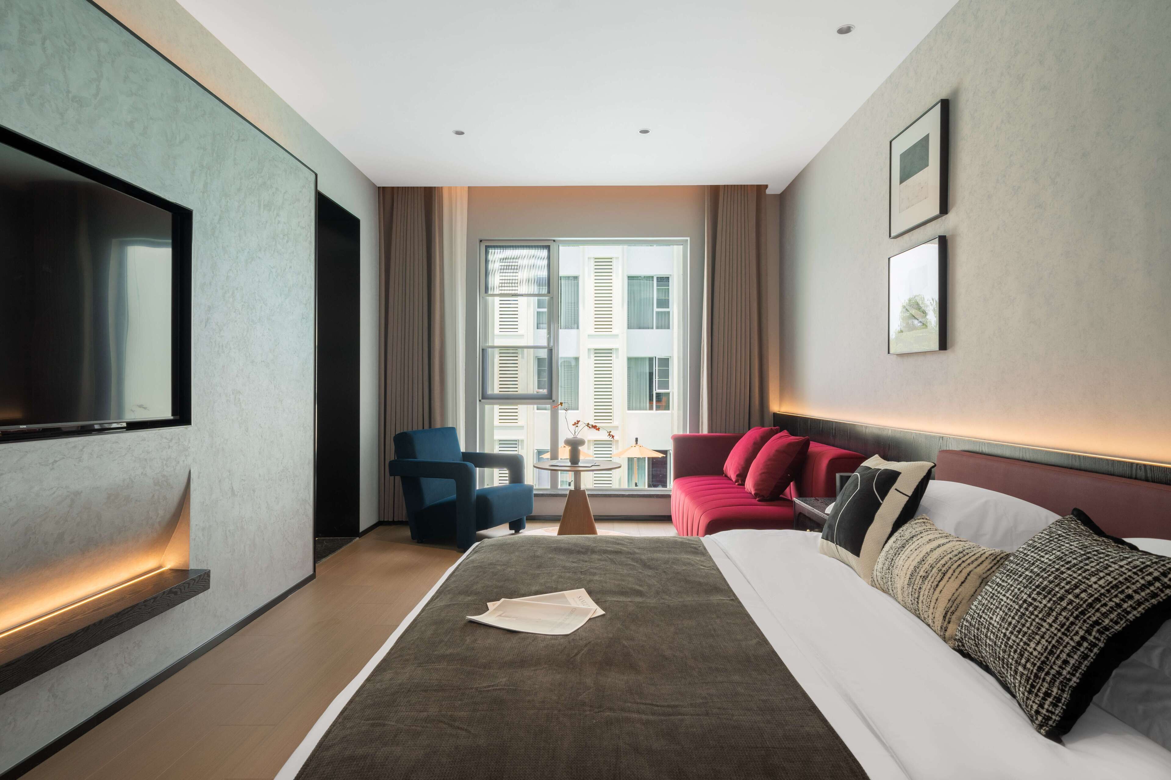 A modern hotel room with a large bed, a flat-screen TV, a red sofa, and a window with a city view.