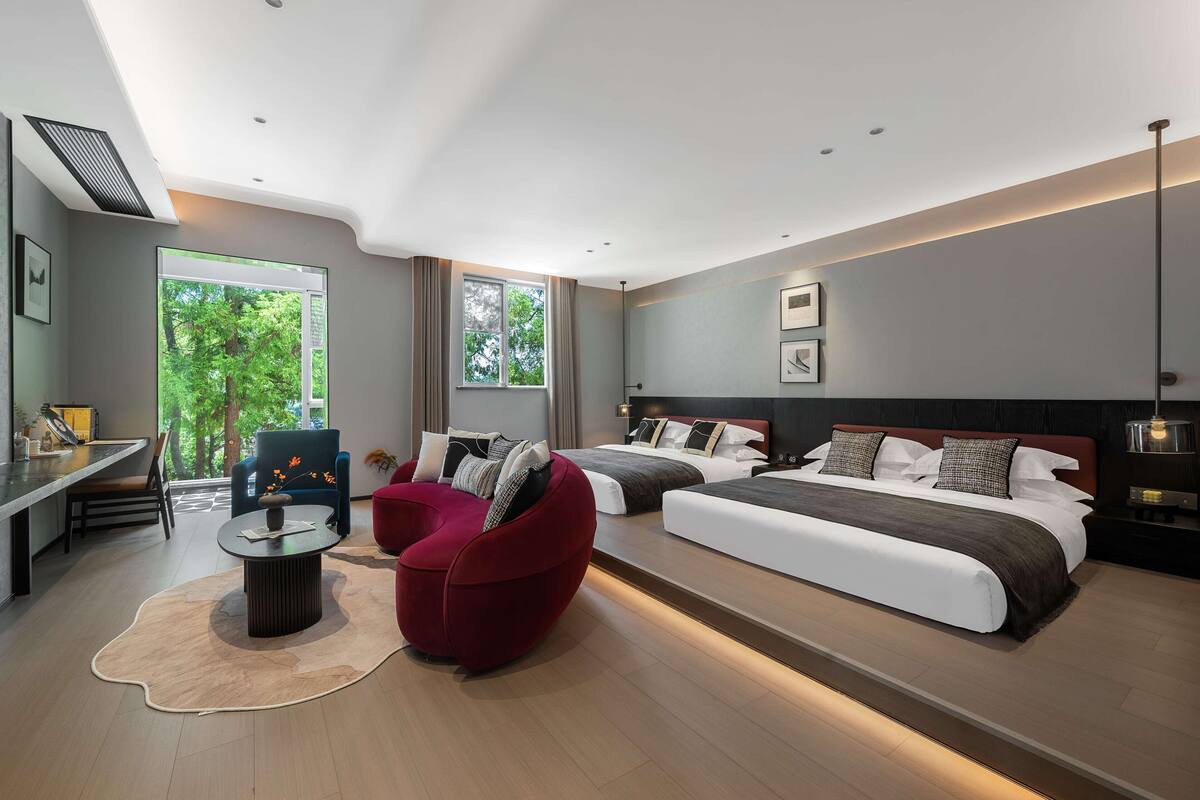 A modern hotel room with a large bed, a red armchair, a dining area, and large windows offering a view of trees.