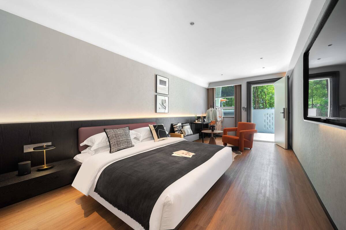A modern hotel room with a large bed, a bedside table with a lamp, a chair, and a view of the outdoors through a sliding door.