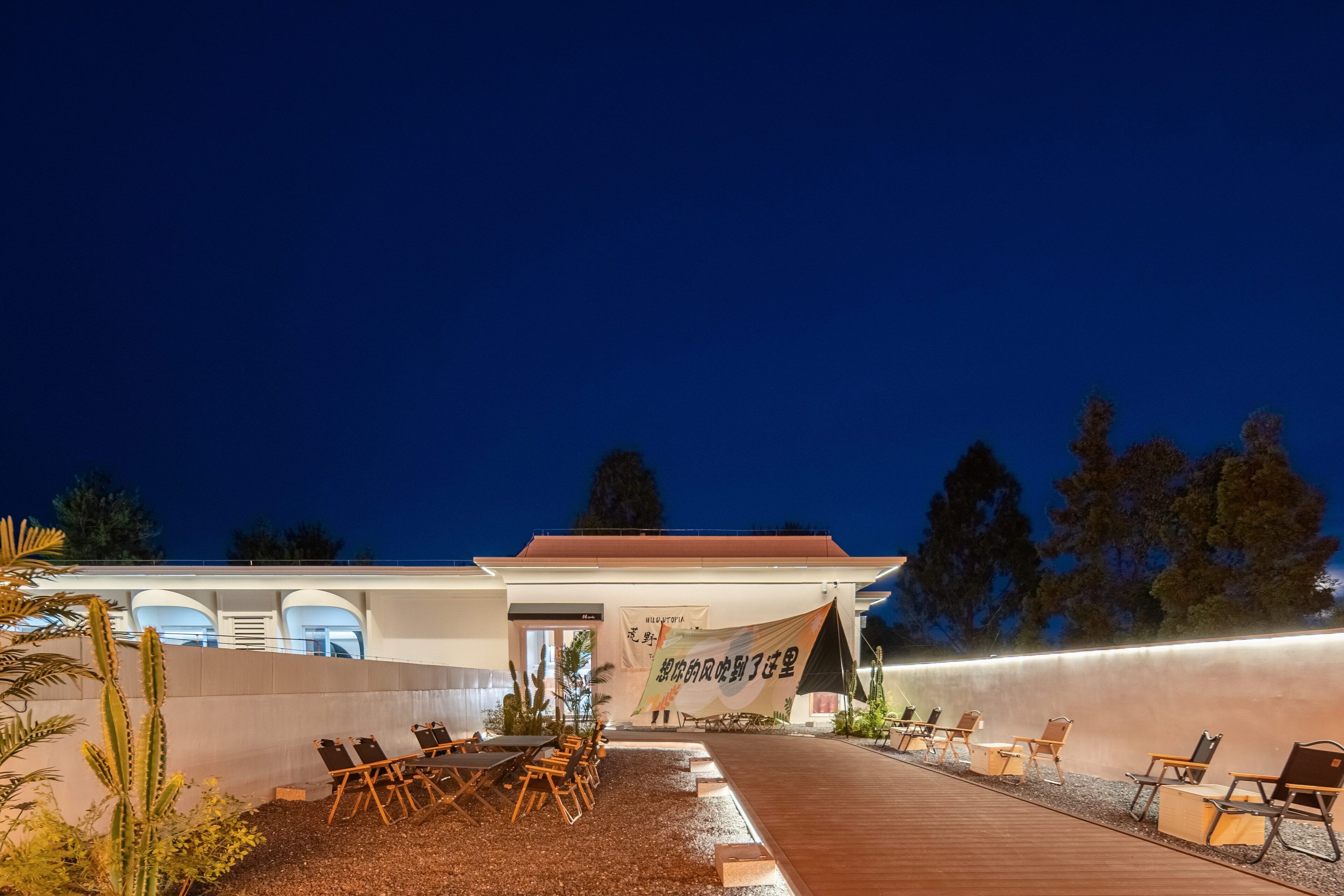 Un área de estar al aire libre con mesas y sillas, un edificio de diseño moderno y un banner con texto.