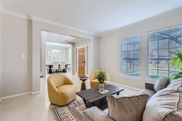 A spacious living room with a sofa, armchair, and coffee table. There is a dining area in the background with a chandelier.