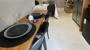 Quarto de hotel moderno com área de refeições, destacando uma mesa de madeira, pratos pretos e um copo. Há uma cama com cabeceira, um sofá e uma televisão.