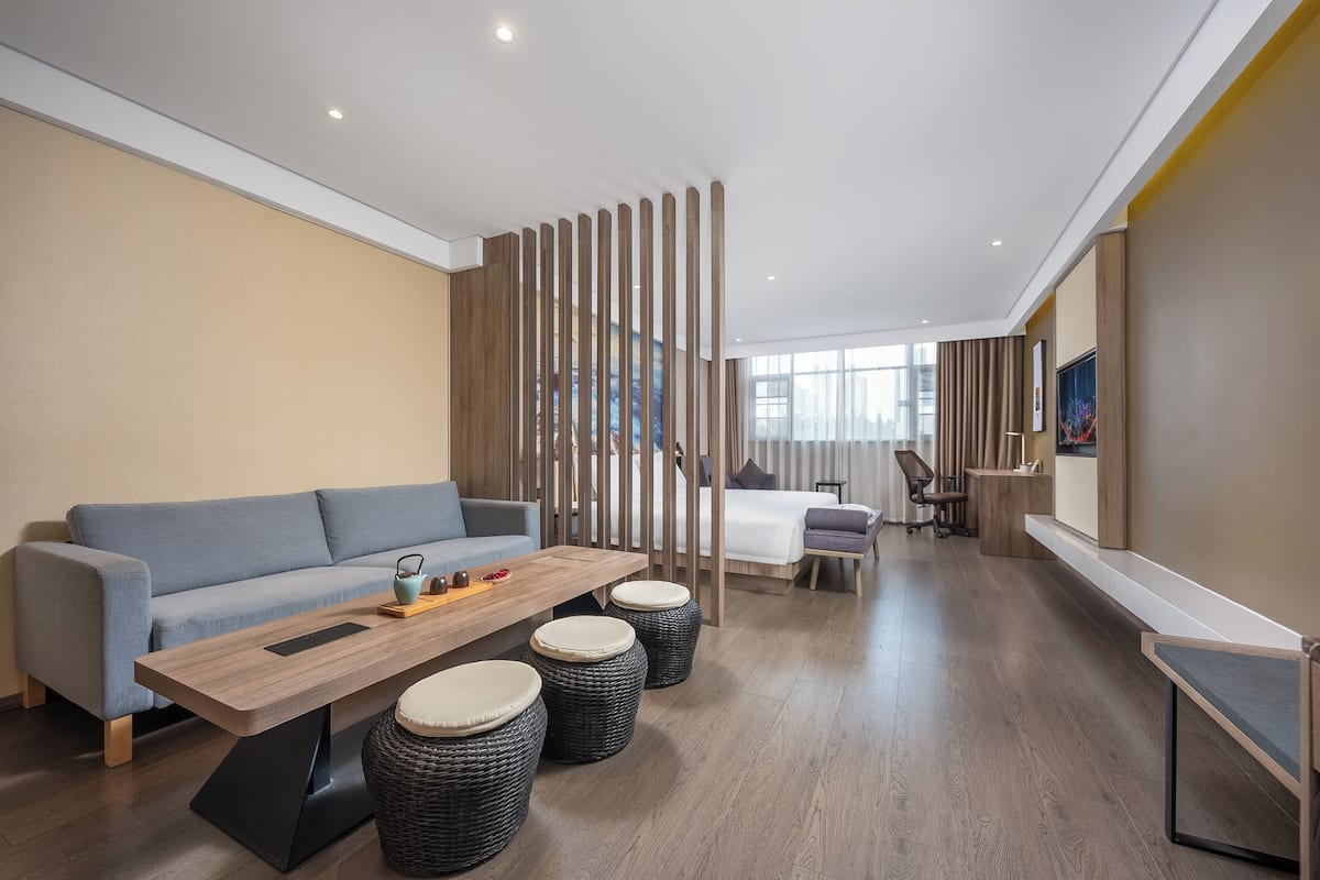 A modern hotel room with a sofa, a wooden table, wicker ottomans, a desk, and a large window.