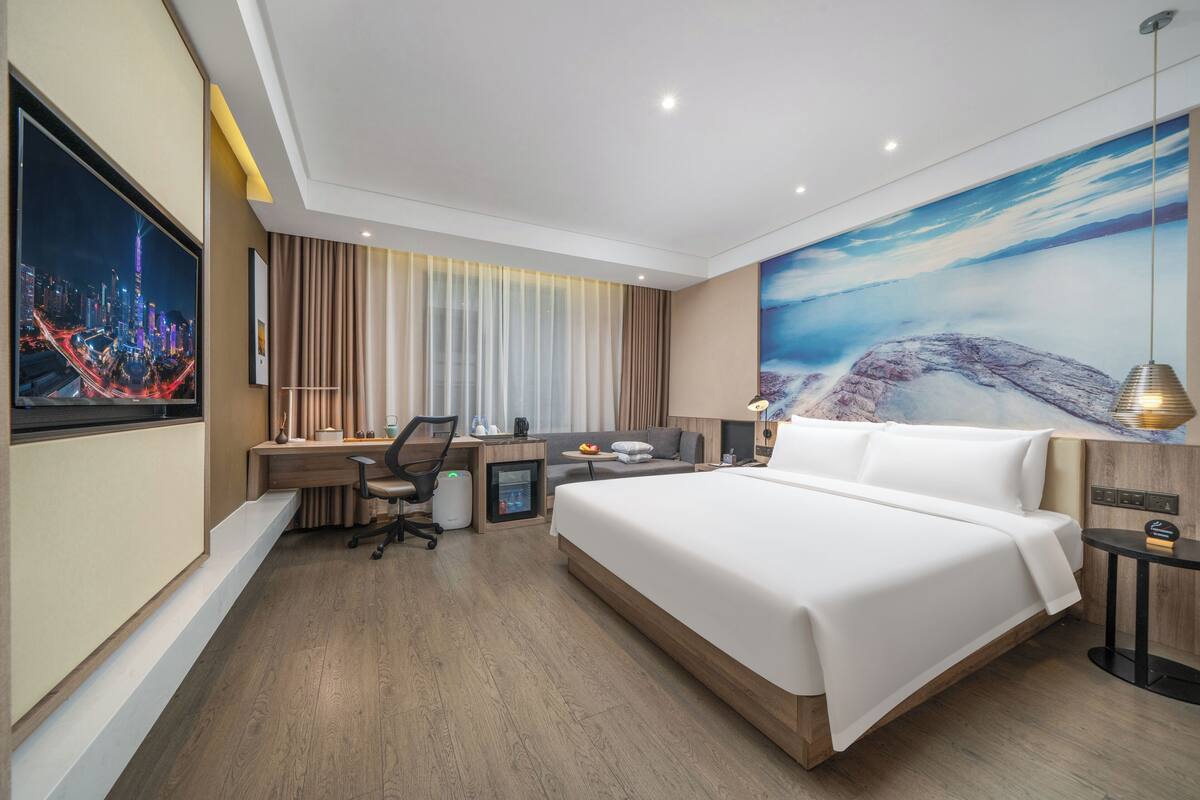 A modern hotel room with a large bed, a desk with a computer, a chair, and a small table with a lamp.