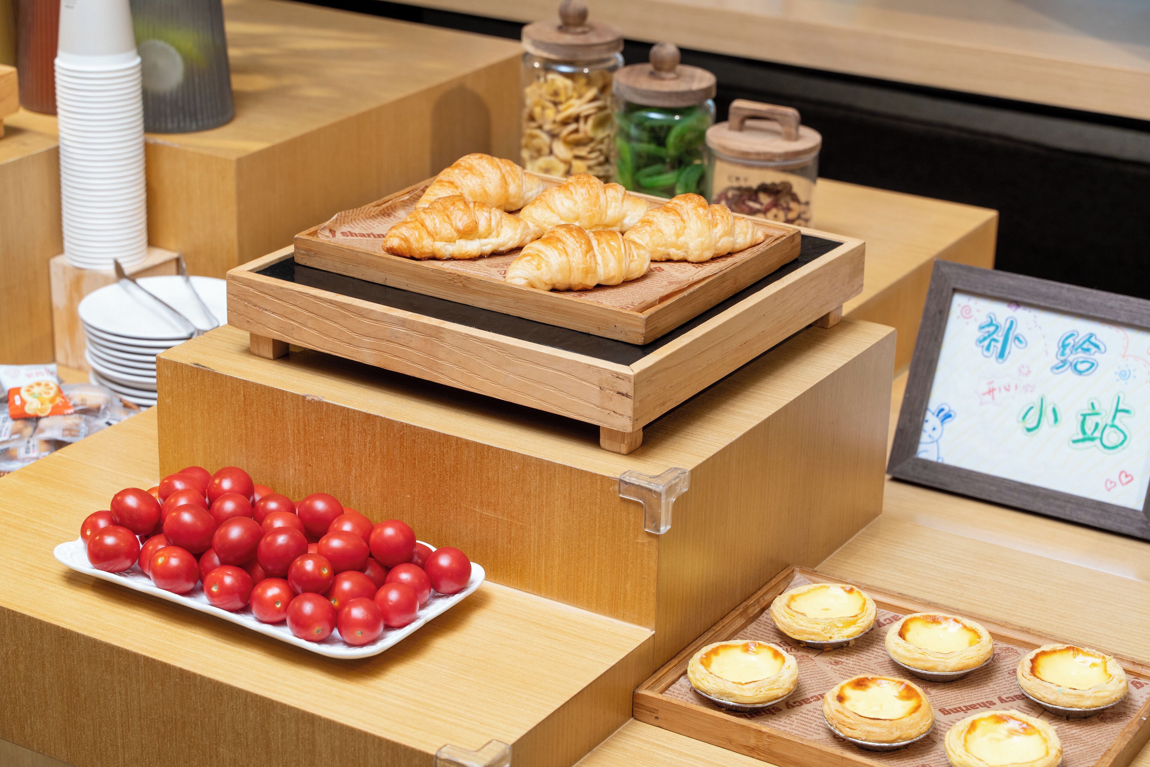 A display of food items including croissants, cherry tomatoes, and custard tarts on a wooden counter.