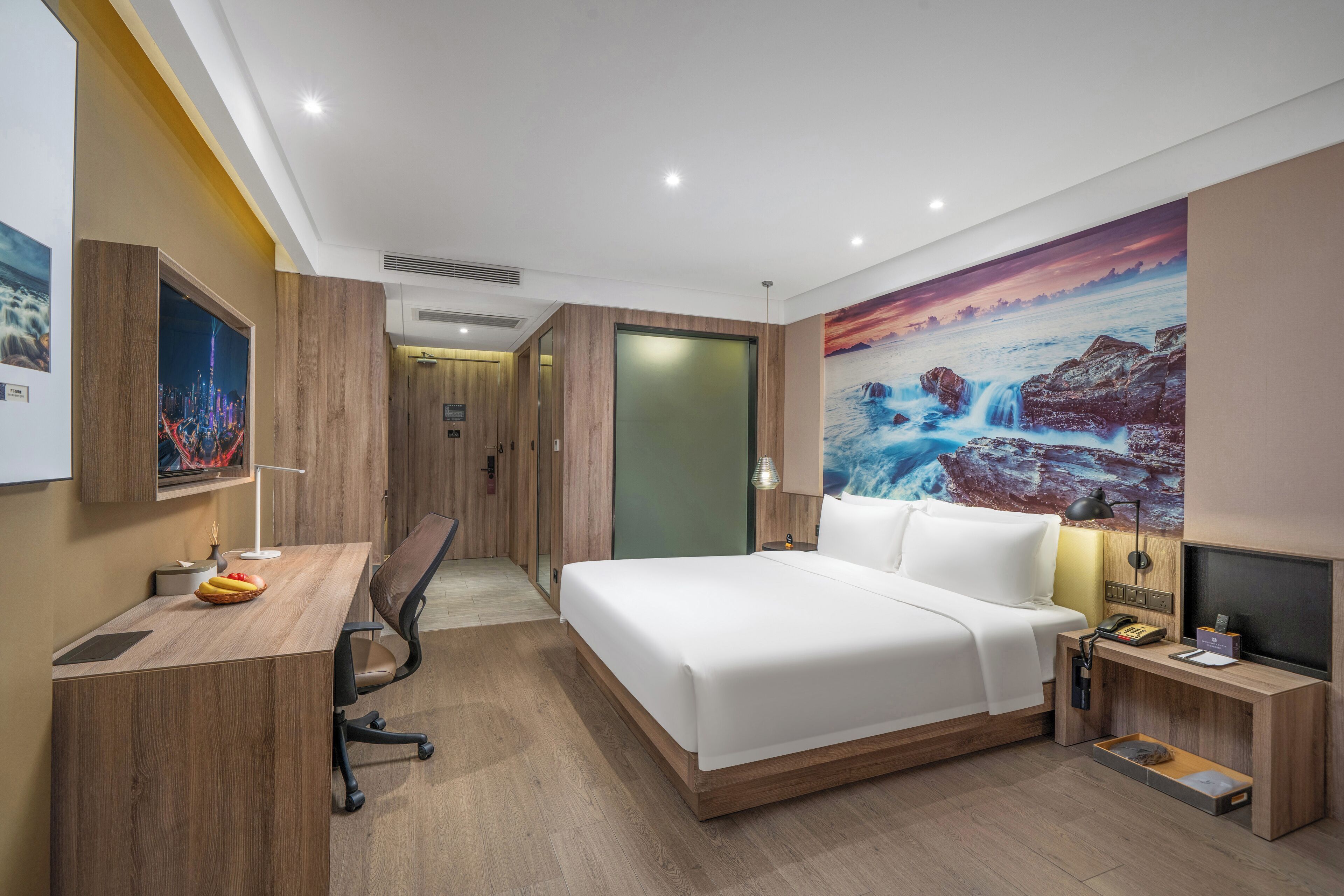 A modern hotel room with a wooden desk, a bed, a nightstand, and a large scenic wall mural.