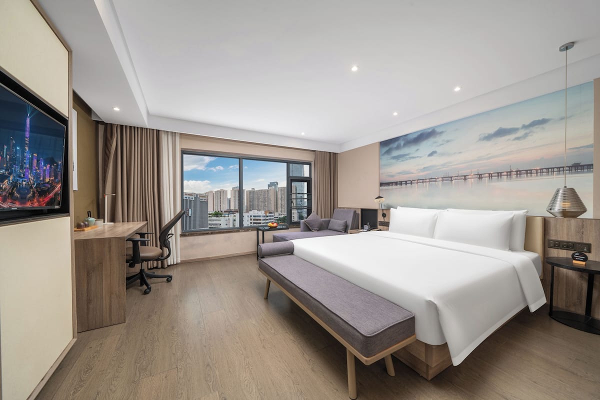 A modern hotel room with a large bed, a desk, a bench, and a city view through the window.