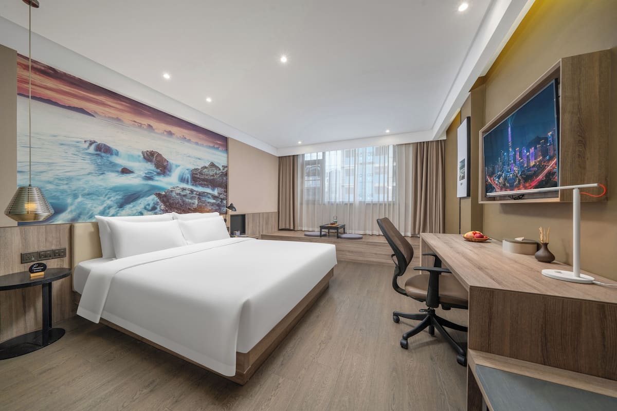 A hotel room with a large bed, a desk, a chair, a TV, and a painting of a seascape.
