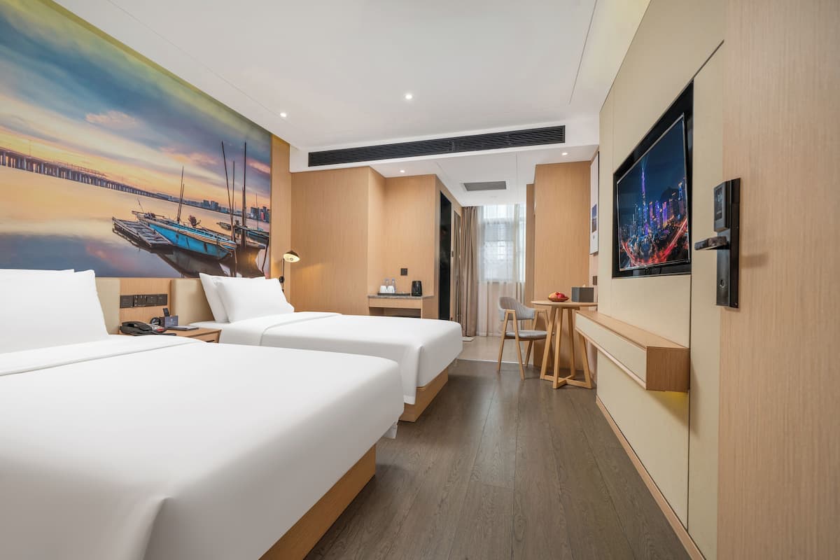 A hotel room with two beds, a desk, a chair, a TV, and a painting of a cityscape.