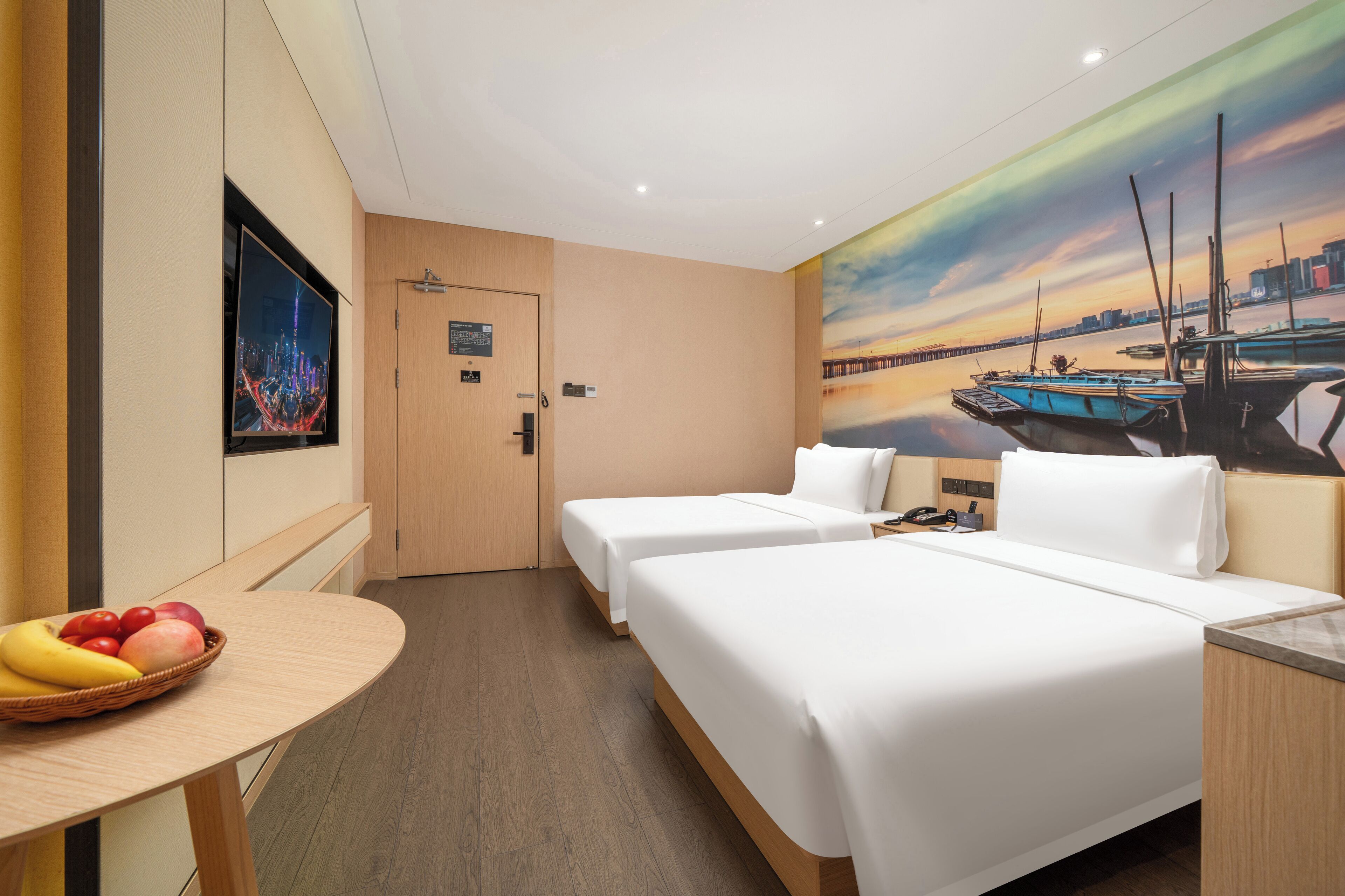 Ein Hotelzimmer mit zwei Betten, einem Esstisch mit Obst, einem Gemälde von Booten und Blick auf eine Stadtlandschaft.