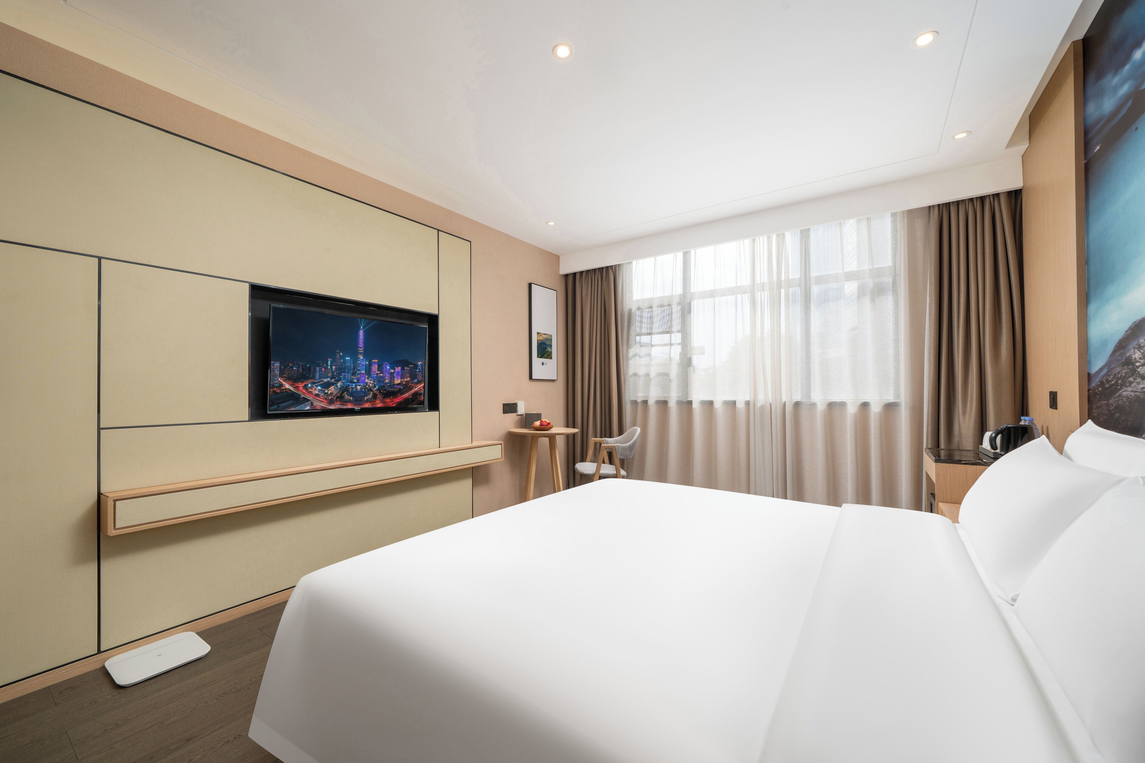 A hotel room with a large bed, a flat-screen TV mounted on the wall, a small table with a chair, and a window with curtains.