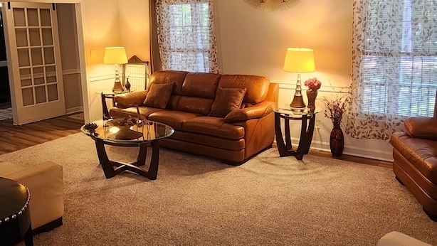 A living room with a brown leather sofa, a white armchair, a glass coffee table, and two side tables with lamps. There is a ceiling fan, a decorative wall clock, and a window with patterned curtains.