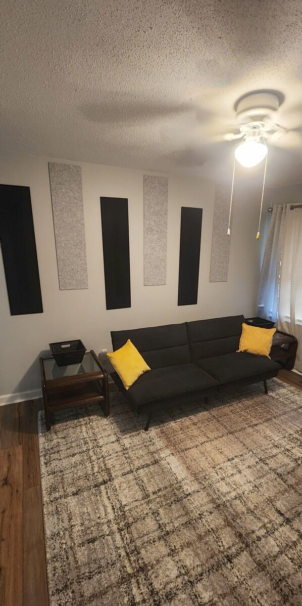 Ein Wohnzimmer mit einem dunklen Sofa, zwei gelben Kissen, einem Glastisch und einem schwarzen Korb.