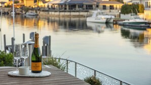 Une bouteille de champagne et deux verres sur une table en bois, près d’un quai, avec des bateaux et des bâtiments en arrière-plan.