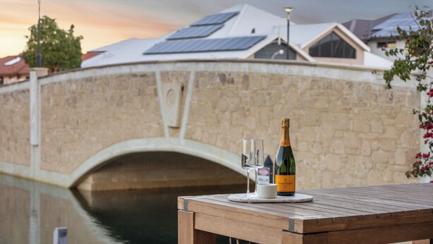 Une table avec une bouteille de champagne et un verre, placée devant un pont de pierre, avec un canal et des bâtiments en arrière-plan.
