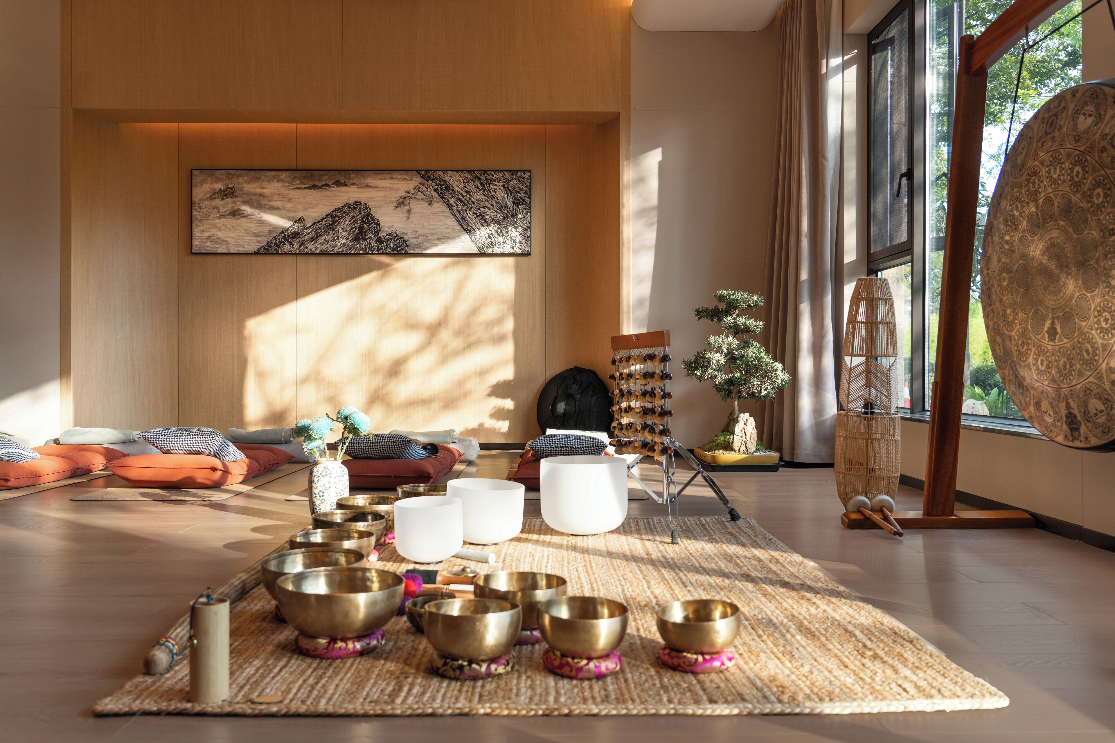 A room with a large window, a wooden floor, and a collection of meditation bowls arranged on a mat.