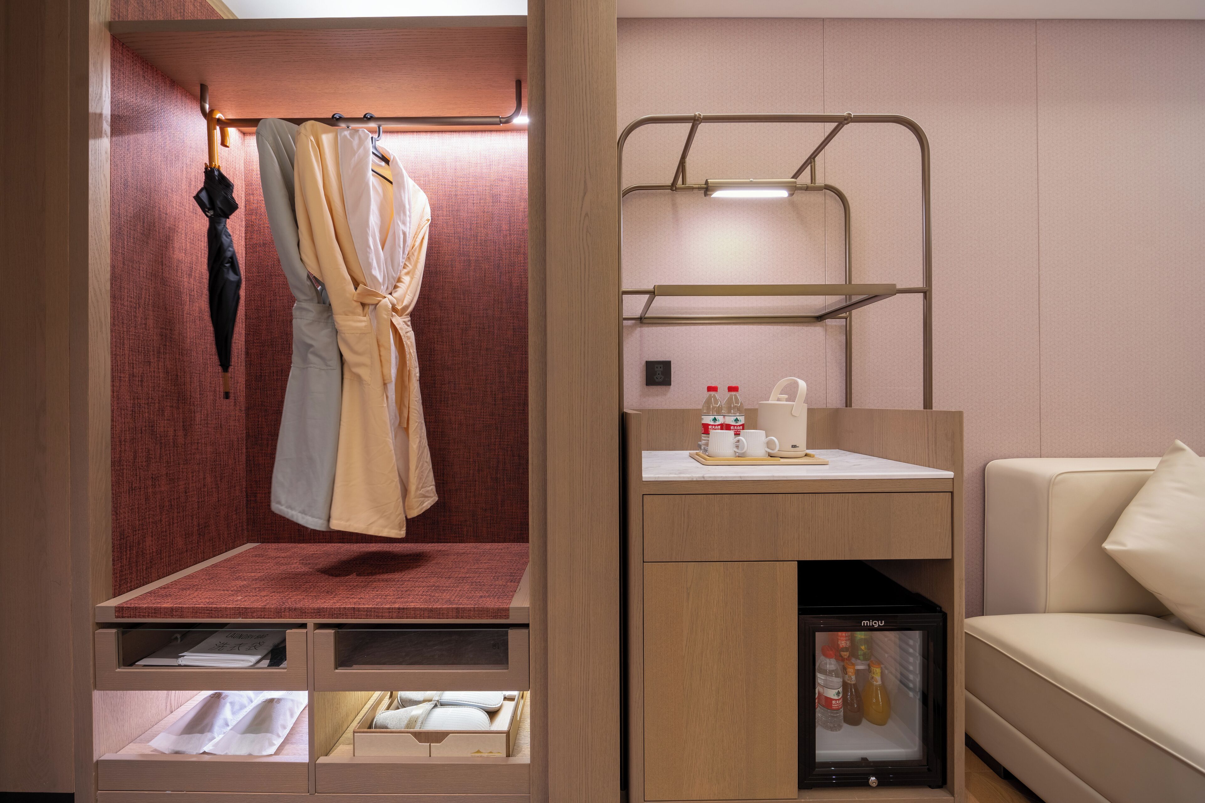 A compact hotel room with a bed, a small seating area with a table and chairs, a minibar, and a coat rack with a robe and umbrella.