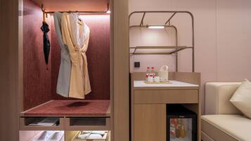 A compact hotel room with a bed, a small seating area with a table and chairs, a minibar, and a coat rack with a robe and umbrella.