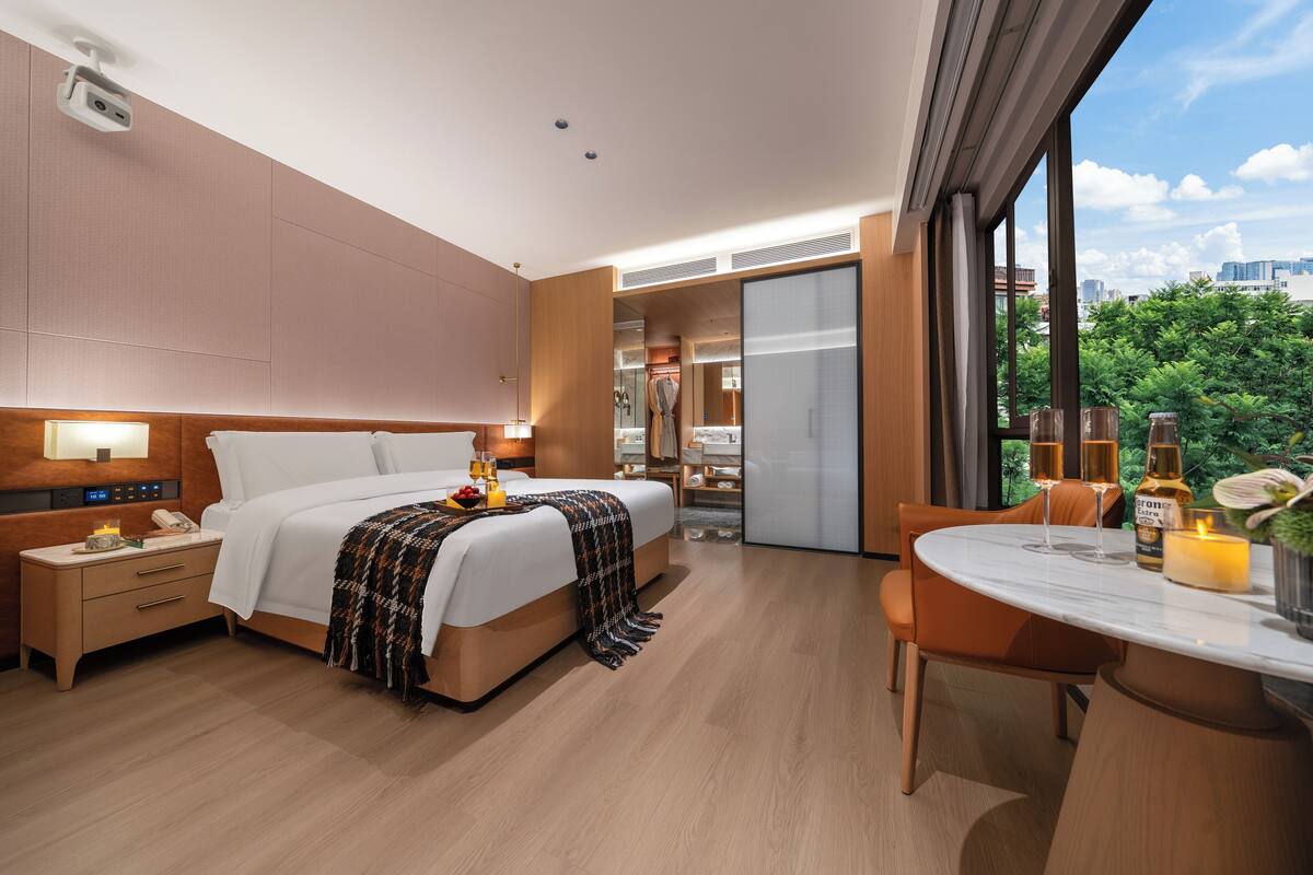A modern hotel room with a large bed, bedside table, and a dining area with a table and chairs.