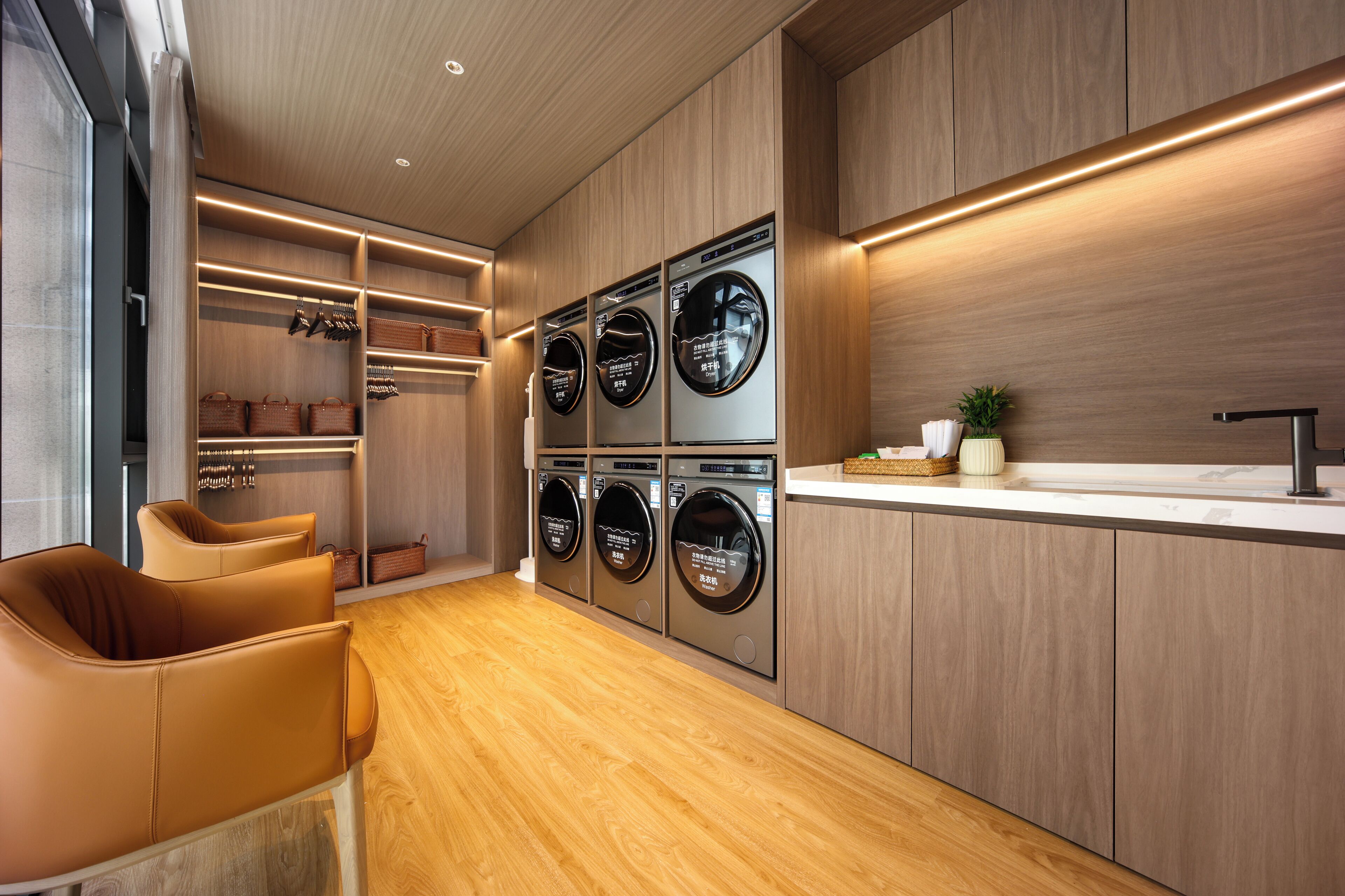 A modern laundry room with built-in washers and dryers, wooden cabinets, and a seating area with chairs.